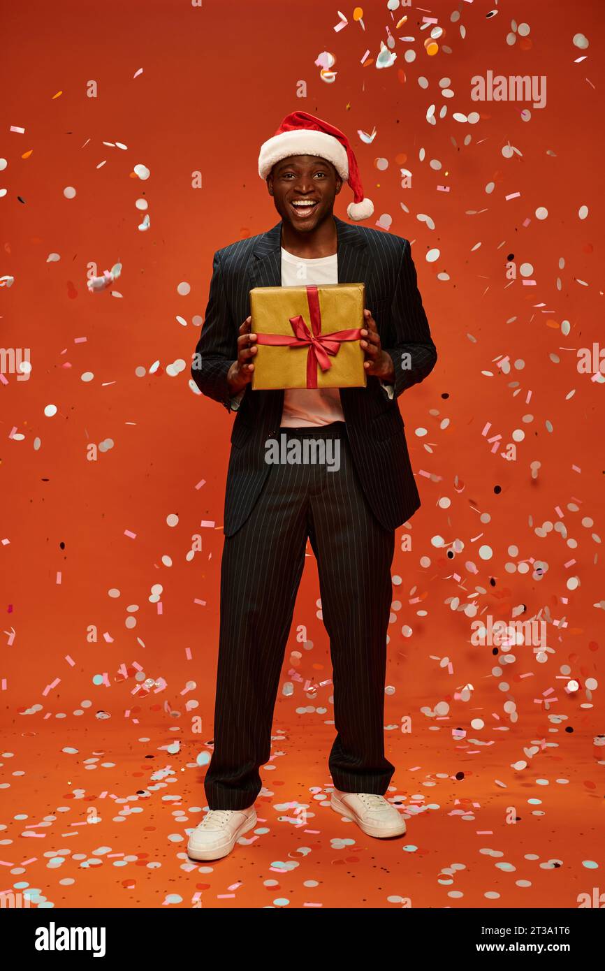 pleased dark skinned man in black suit and santa hat standing with gift box under confetti on red Stock Photo