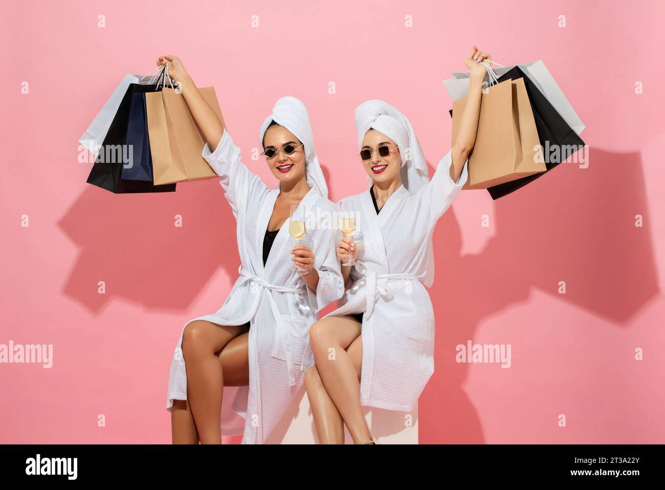 Two woman friends wearing spa bathrobes holding shopping bags and drinks in pink color isolated background studio shot Stock Photo