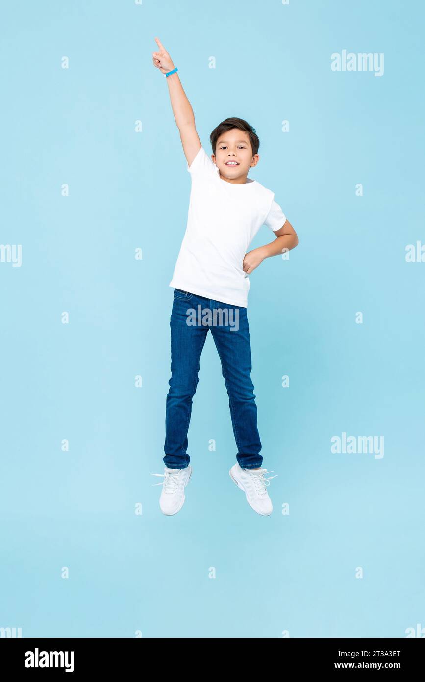 Young mixed race Asian kid boy jumping with hand pointing up in the air against light blue color isolated background studio shot Stock Photo