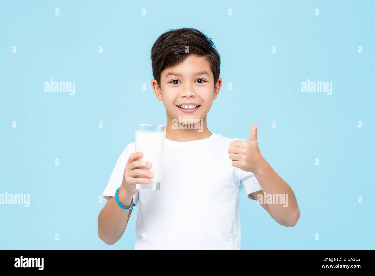 Happy smiling mixed race Asian kid boy holding a glass of fresh milk and giving thumbs up in isolated light blue color background studio shot Stock Photo