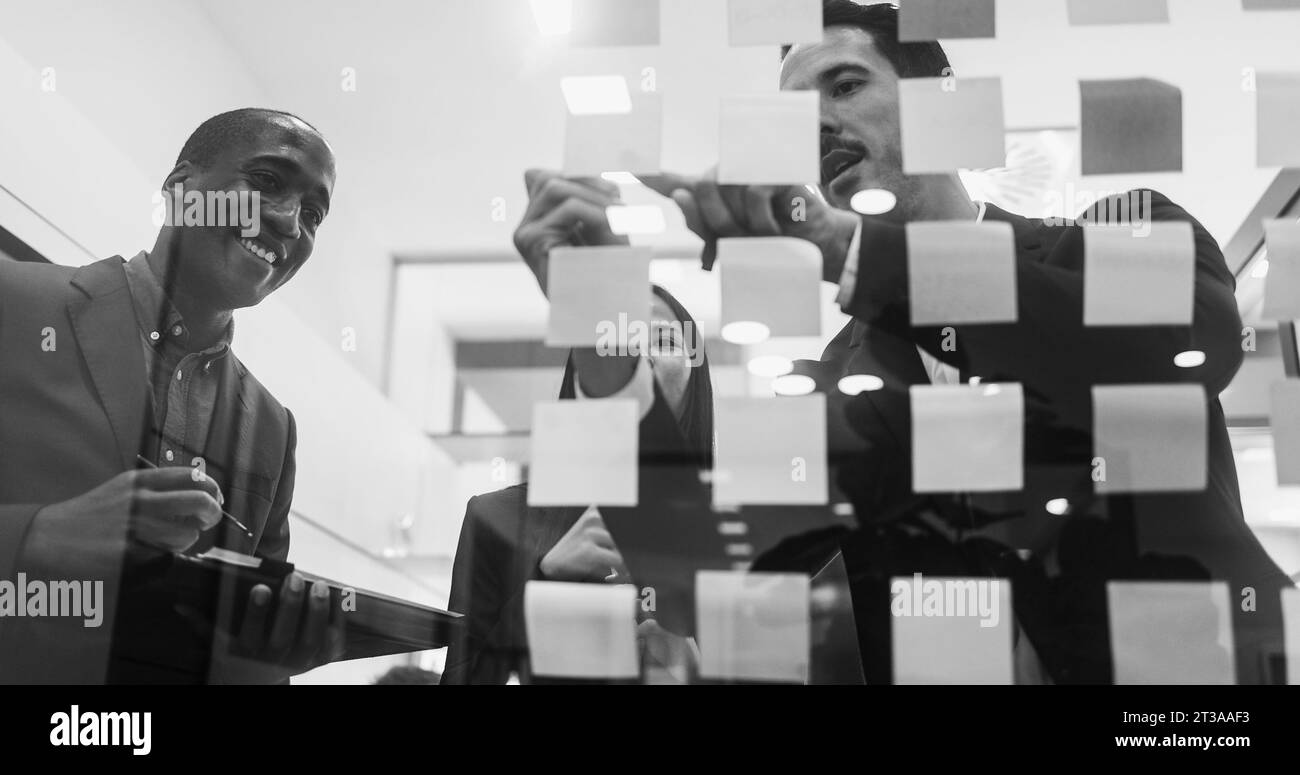 Multiracial colleagues working inside business office with glass walls - Entrepreneurs people sharing ideas at teamwork meeting - Black and white edit Stock Photo