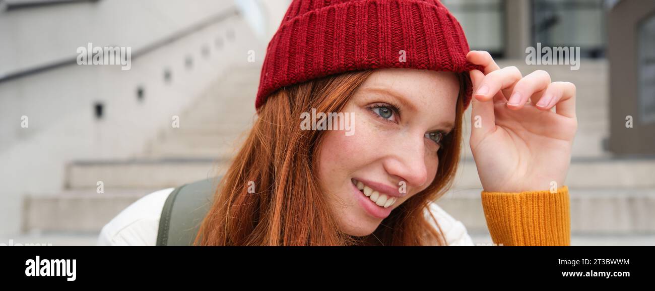 Stylish redhead girl in warm red hat, smiling relaxed, sitting with backpack on stairs near building, waits for someone outdoors Stock Photo