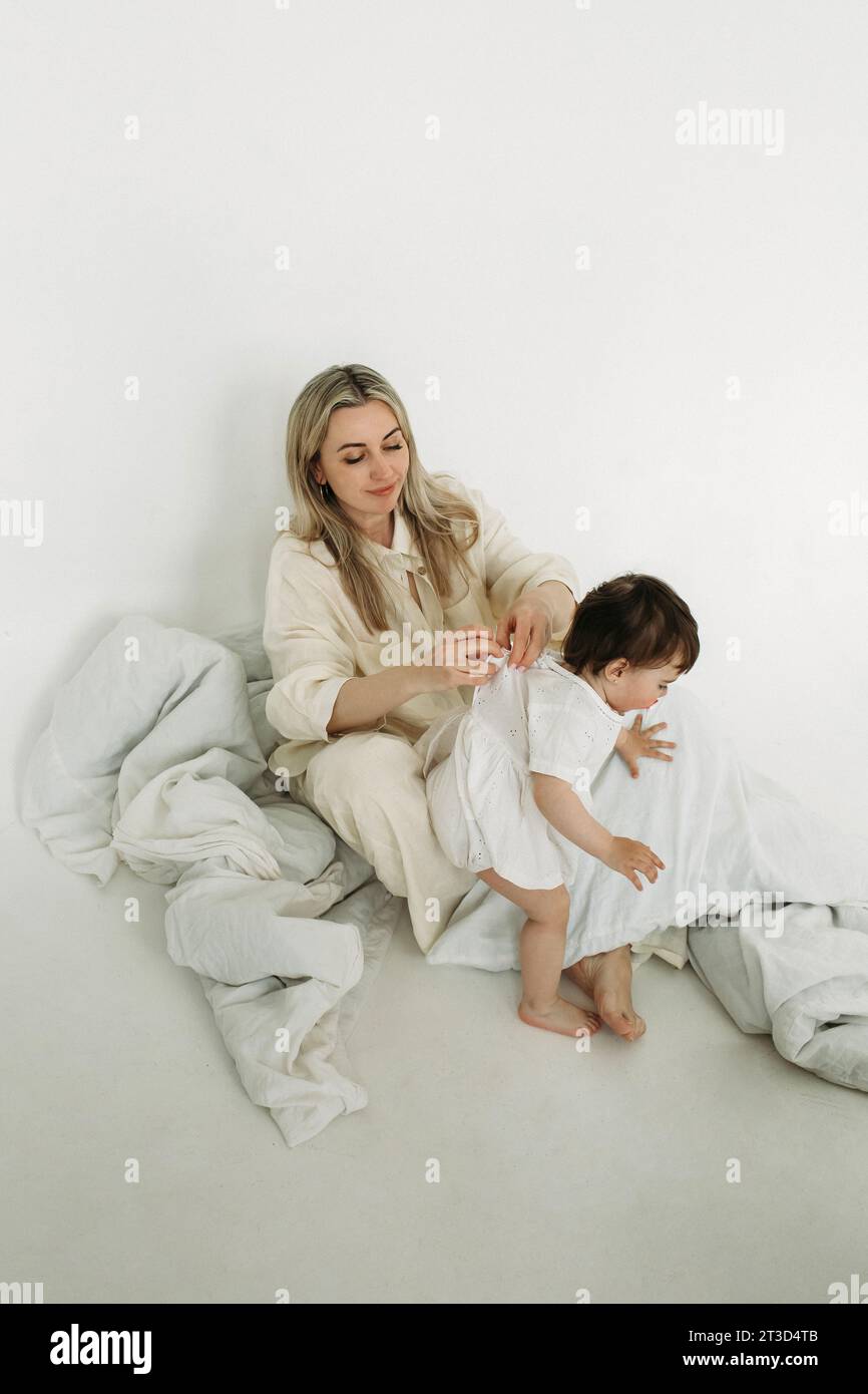 Mom dresses - fastens her little daughter's dress after sleep. Vertical frame. Stock Photo