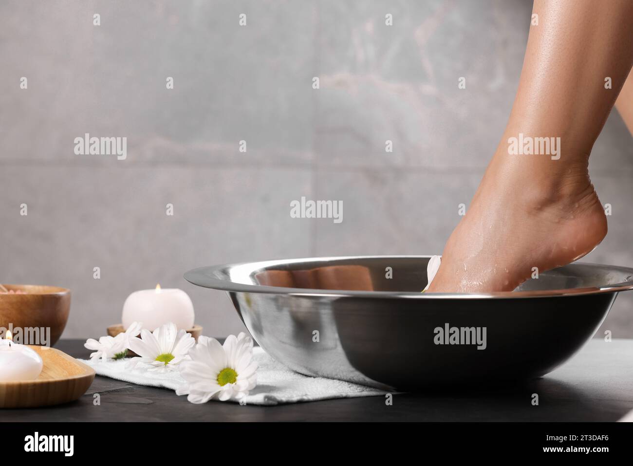 Woman soaking her foot in bowl with water on dark surface, closeup