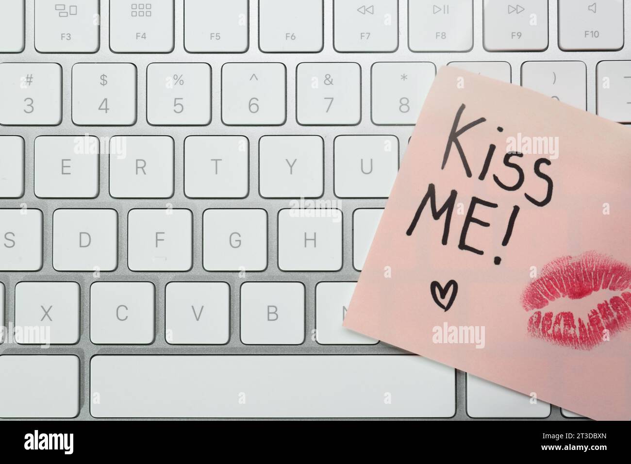 Sticky note with phrase Kiss Me and lipstick mark on keyboard, top view