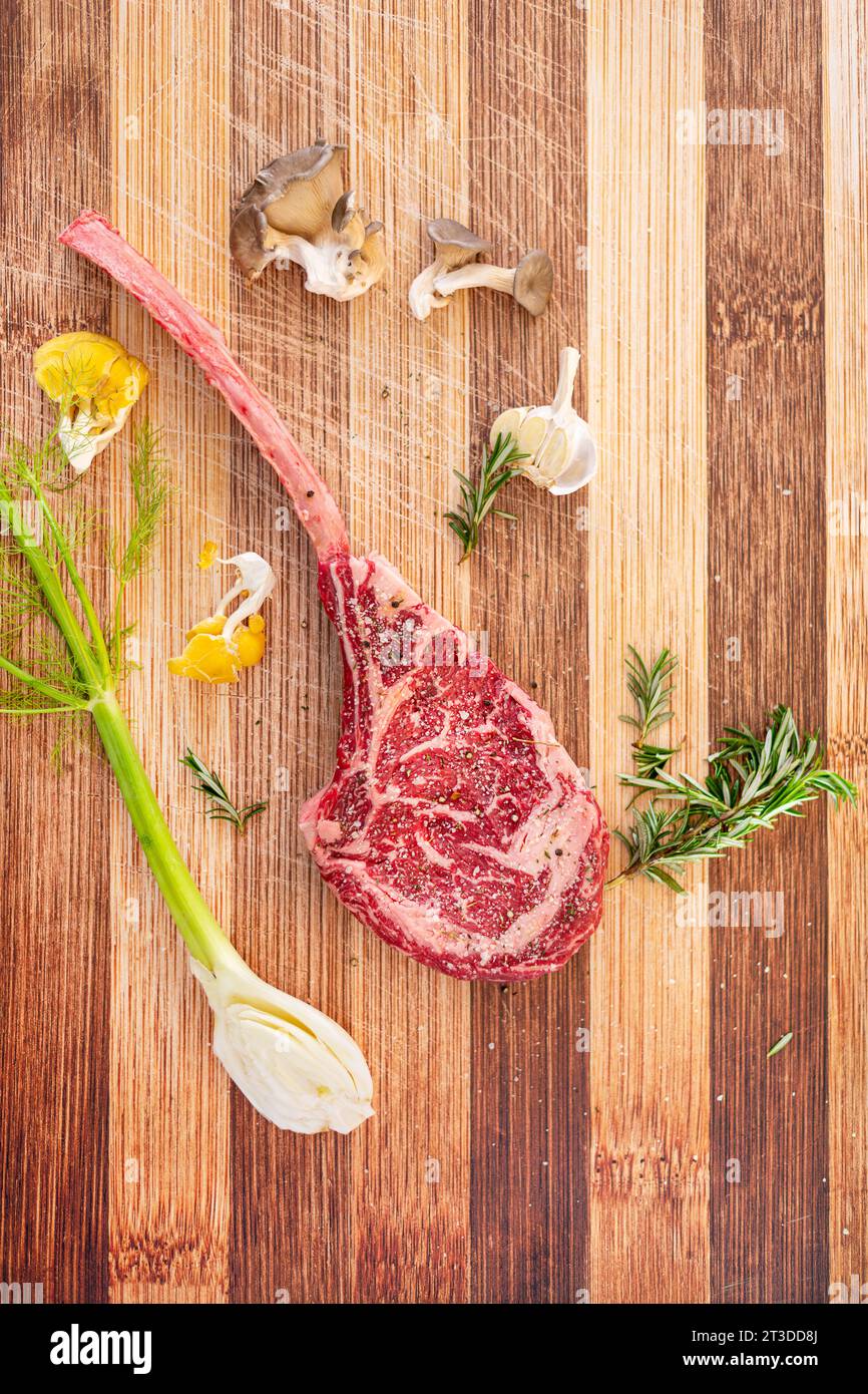 A well marbled tomahawk stak on the butchers block with fresh ingredients of rosemary, oyster mushrooms, fennel and peppercorns. Stock Photo