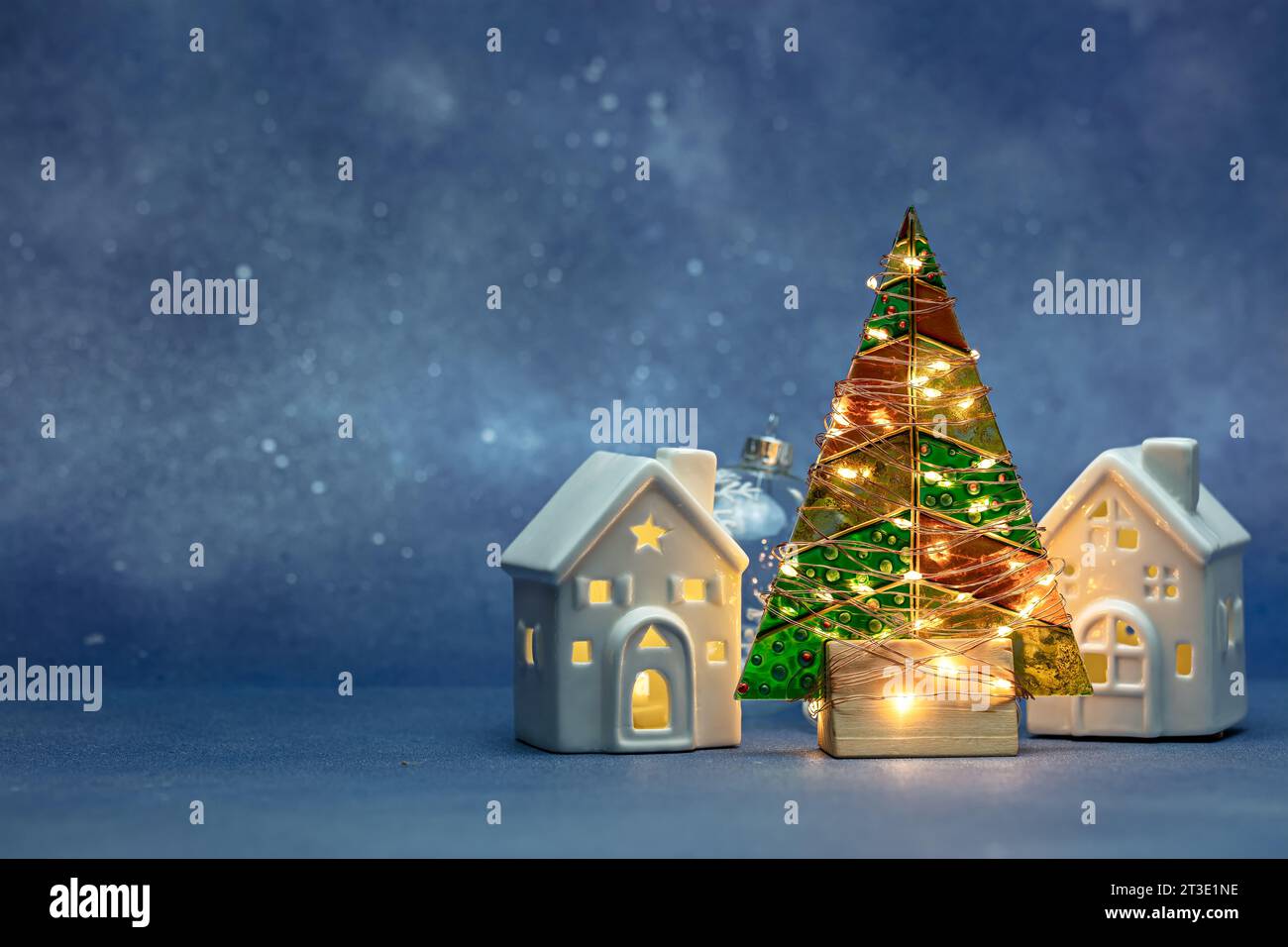 glass christmas tree with glowing holiday lights and two illuminated toy homes on blurred blue background. Stock Photo