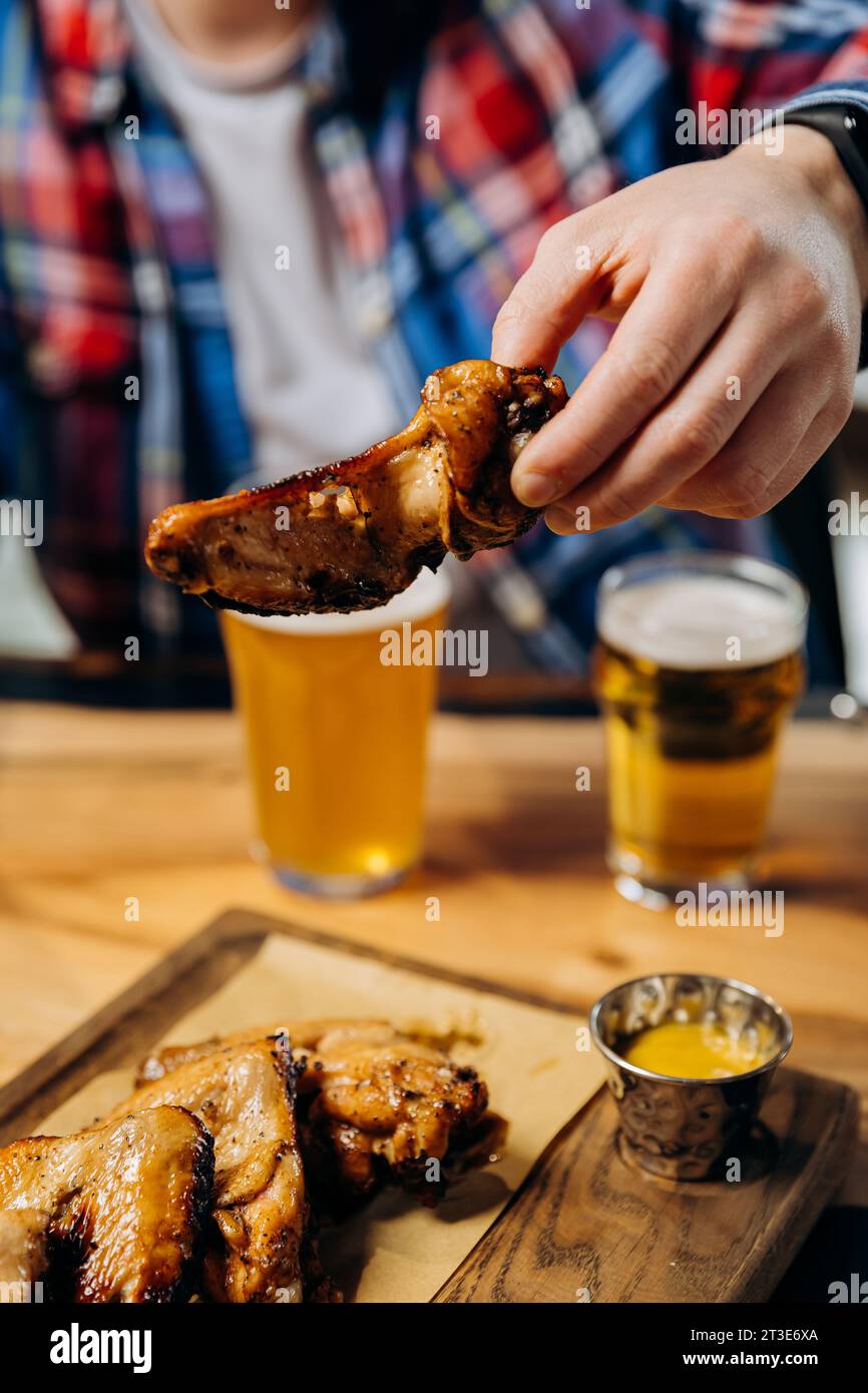 Male hand holding spicy grilled BBQ chicken wings and eating with beer in pub Stock Photo