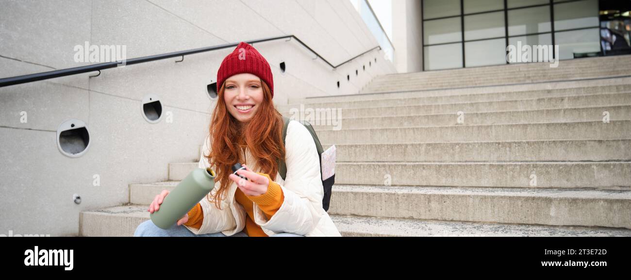 Happy redhead woman, tourist with thermos, drinks her hot tea, coffee from travel flask, restests during her travelling in city and sightseeing Stock Photo