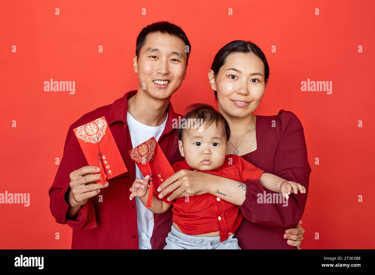 Waist up portrait of young Chinese family with baby boy standing against red and smiling at camera holding red envelopes Have overflowing abundance every year Stock Photo