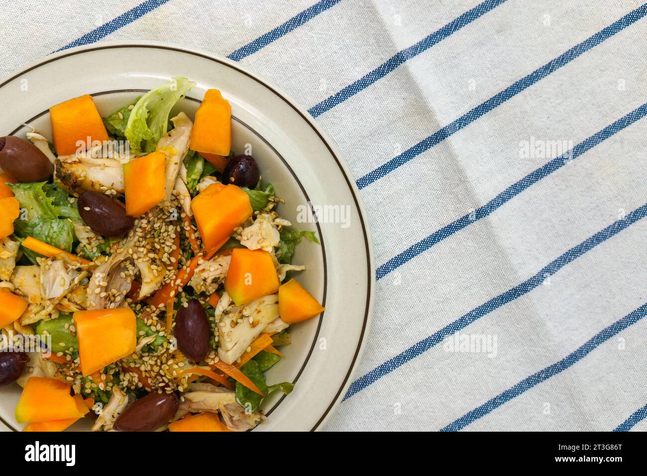 Fresh chicken salad with kalamata olives, cantaloupe melon and sesame seeds. Top view on white kitchen towel with blue stripes. Stock Photo