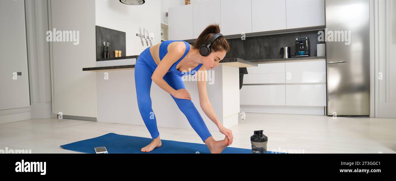 Image of young fit woman doing fitness workout from home, stretching her legs, warm-up before aerobics training, listening music in wireless headphone Stock Photo