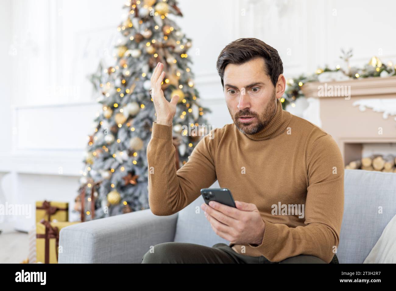Shocked young man looking at phone screen and reading bad news, sitting at home on sofa near Christmas tree on holidays and gesturing angrily with hands. Stock Photo