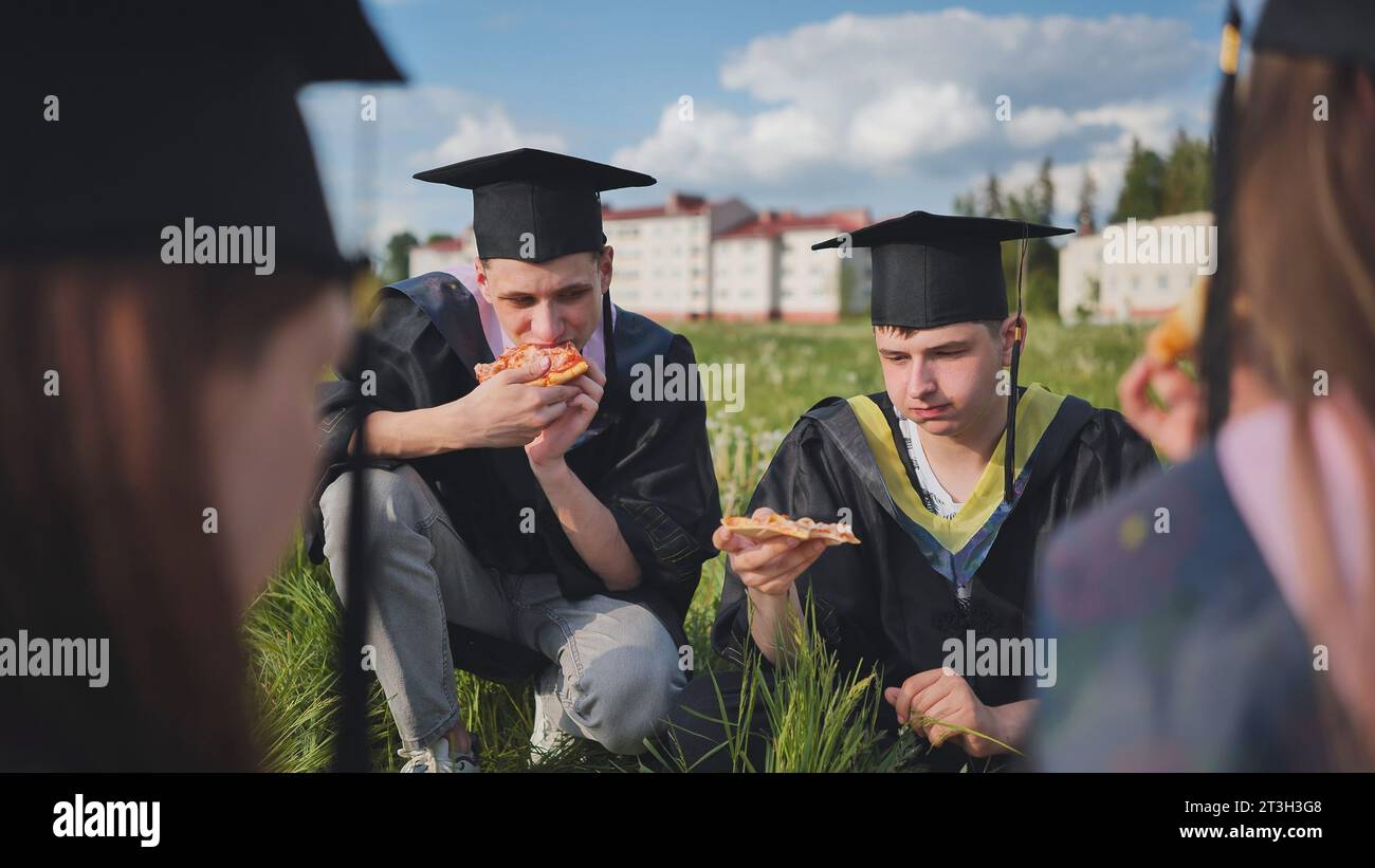 Graduates in black suits eating pizza in a city meadow Stock Photo - Alamy