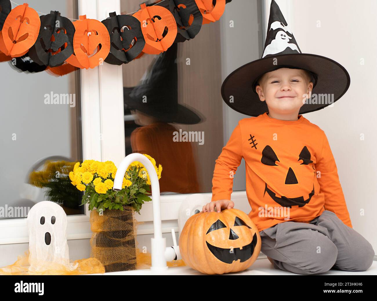 Halloween concept. A small, cheerful, handsome boy in a wizard's hat and an orange sweater sits on a table in the kitchen against the background of a Stock Photo