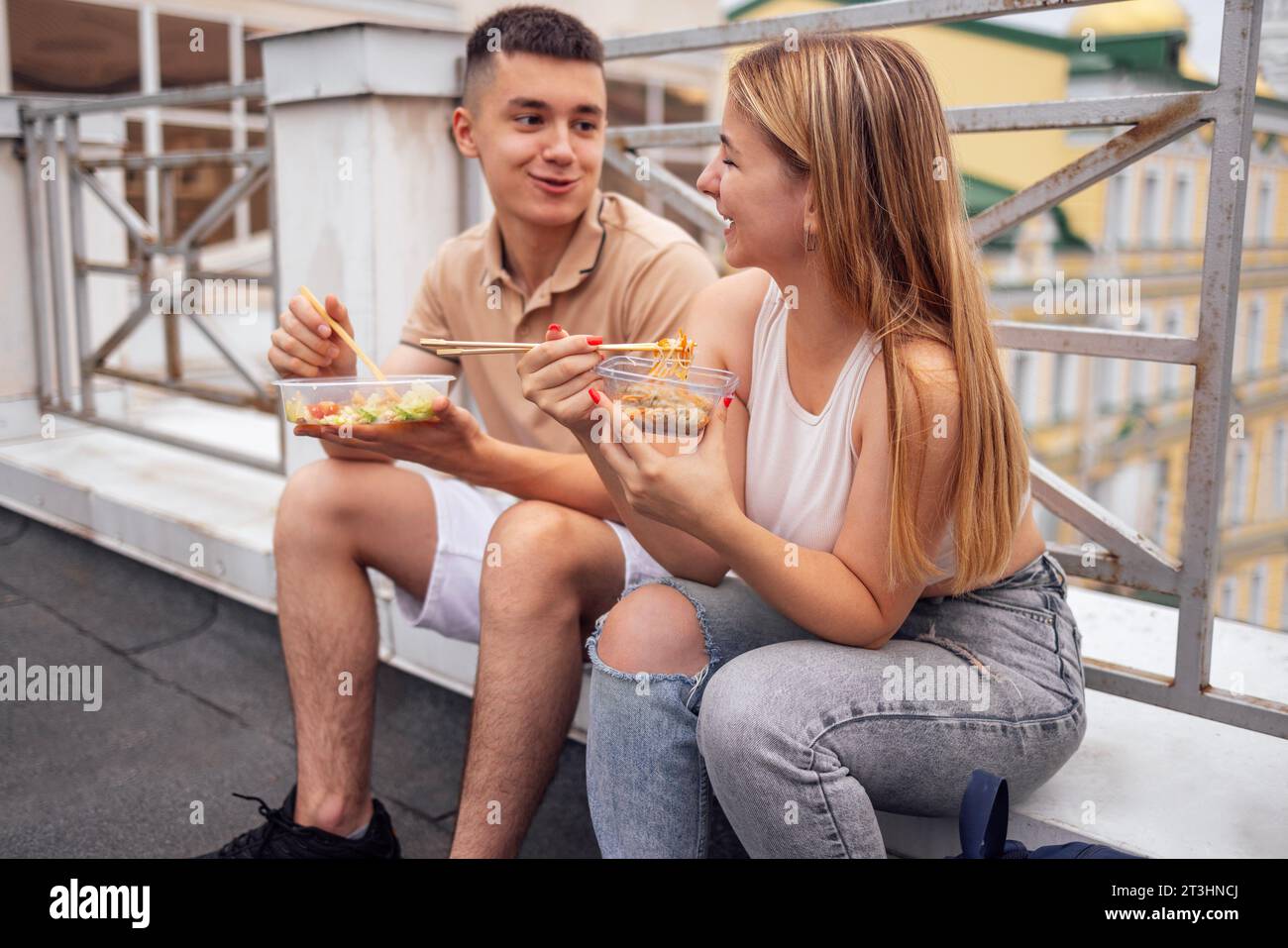 Two teenagers are sitting on the parapet on the roof of the house and eating Asian food. Young couple snack on fast food and have fun together. Girls Stock Photo