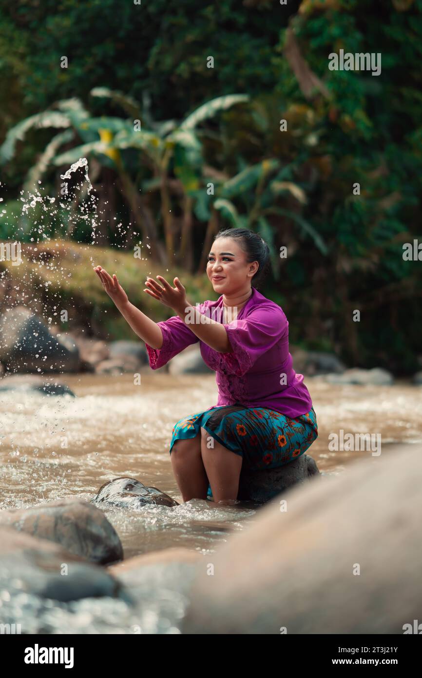 Asian woman playing with dirty water from a dirty river while wearing a purple dress and green skirt near the river Stock Photo