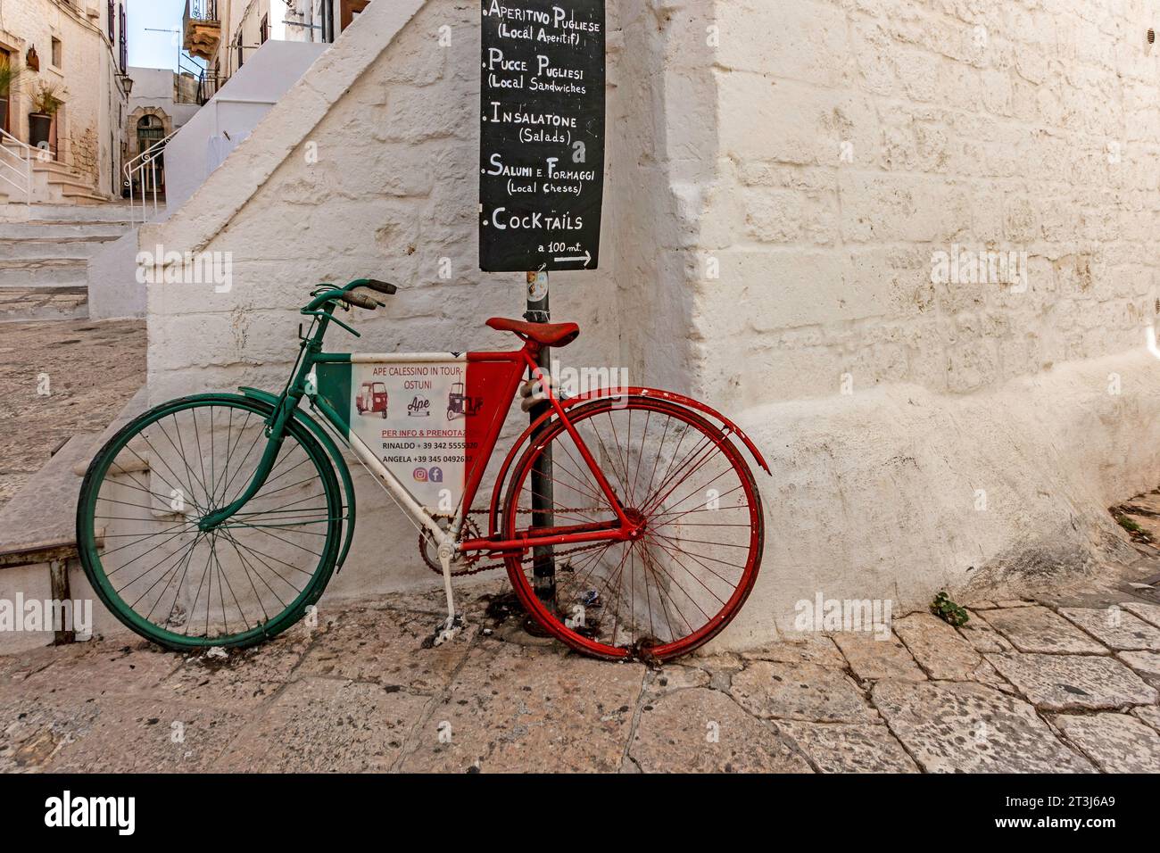A colourful scene in Ostuni, Italy, a bicycle carrying an advertisement for TucTuc motor tours  under a sign for a local cafe. Stock Photo
