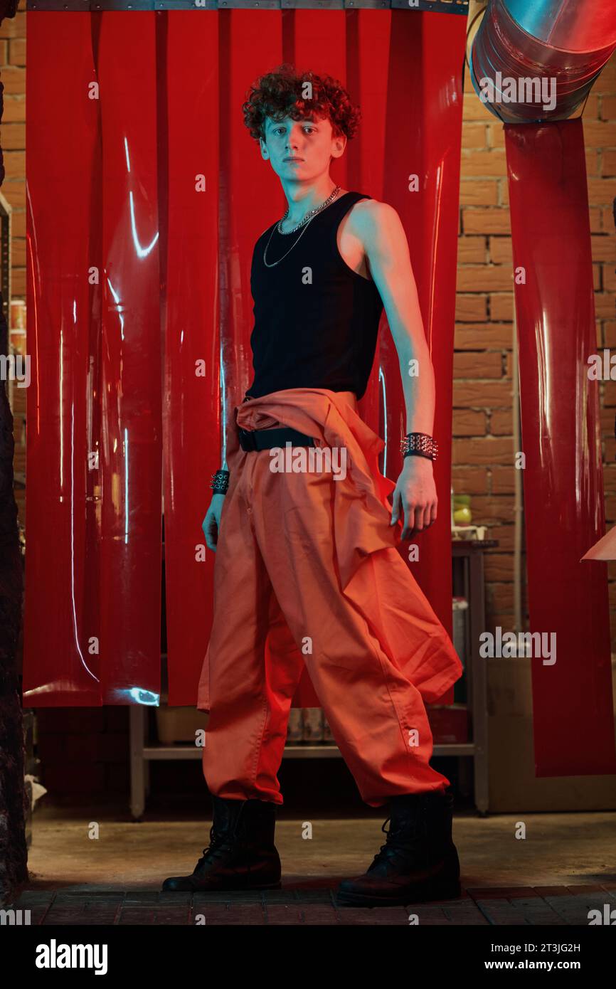 Long shot of young stylish man in pants and singlet looking at you while standing in front of camera against red curtains in cyberpunk bar Stock Photo