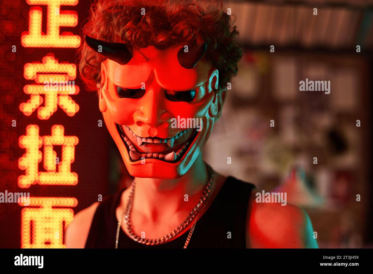 Young man in devil mask standing against hieroglyph illumination meaning best ramen and looking at camera in cyberpunk club, bar or cafe Stock Photo