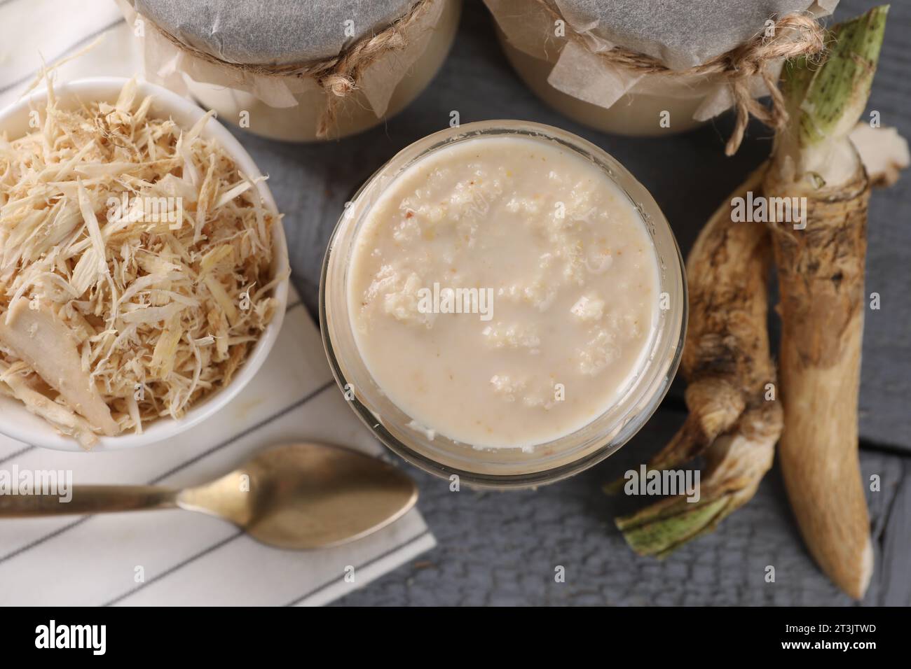 Spicy horseradish sauce in jars, roots and spoon on grey wooden table