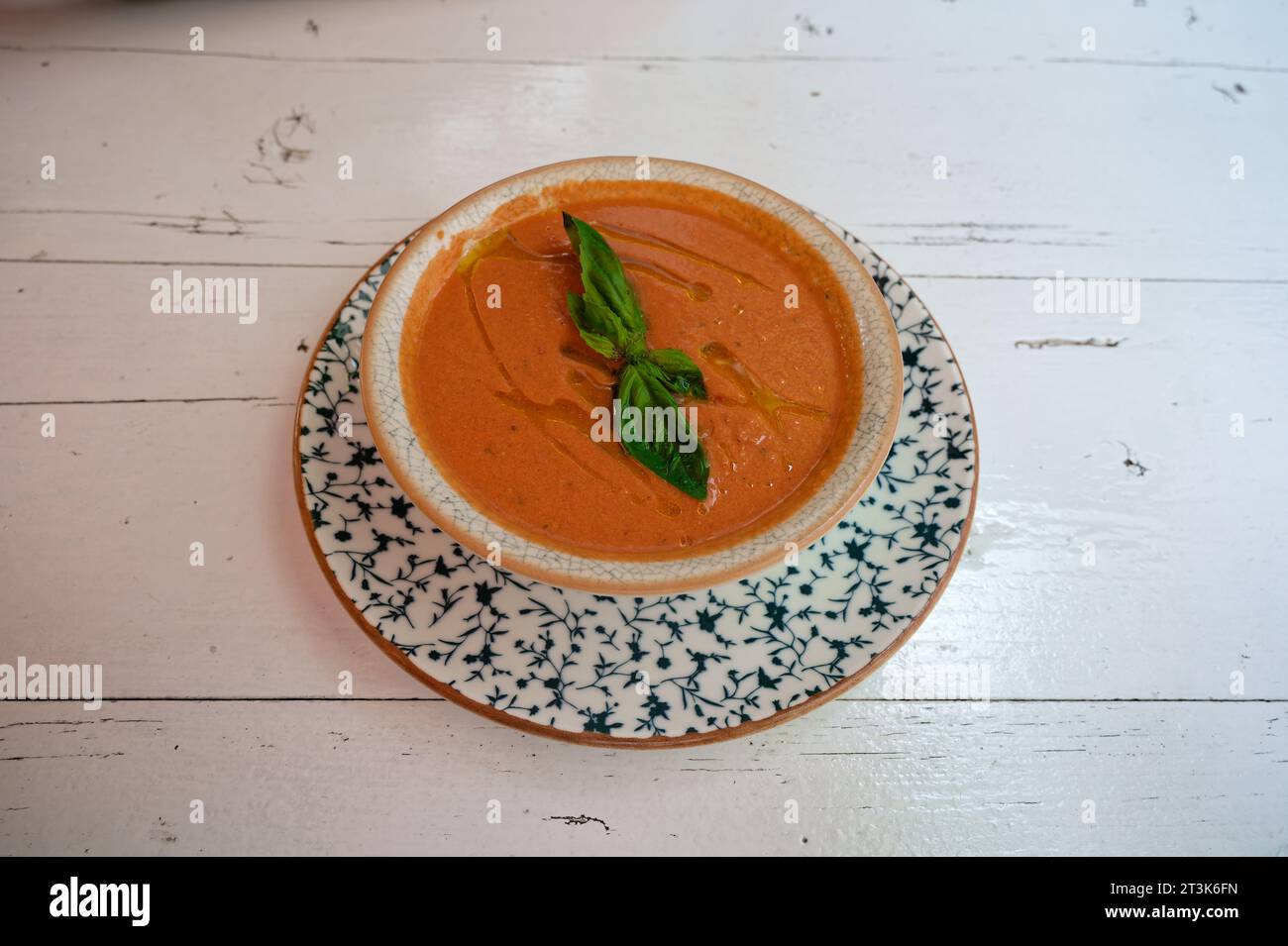 Gazpacho with vibrant orange and green colors, served in a rustic bowl on a beautiful blue plate atop a white, weathered table. Stock Photo