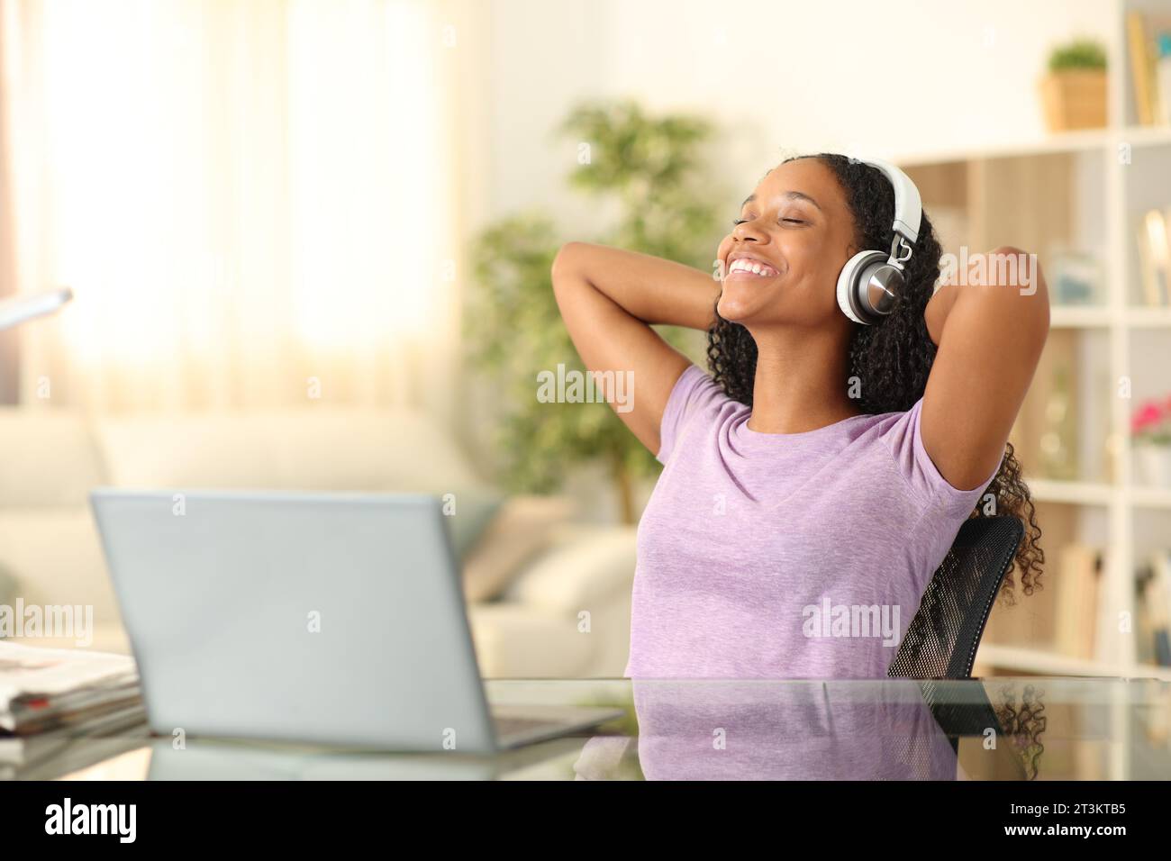 Happy black woman listening music with headphone and laptop relaxing sitting on a chair at home Stock Photo