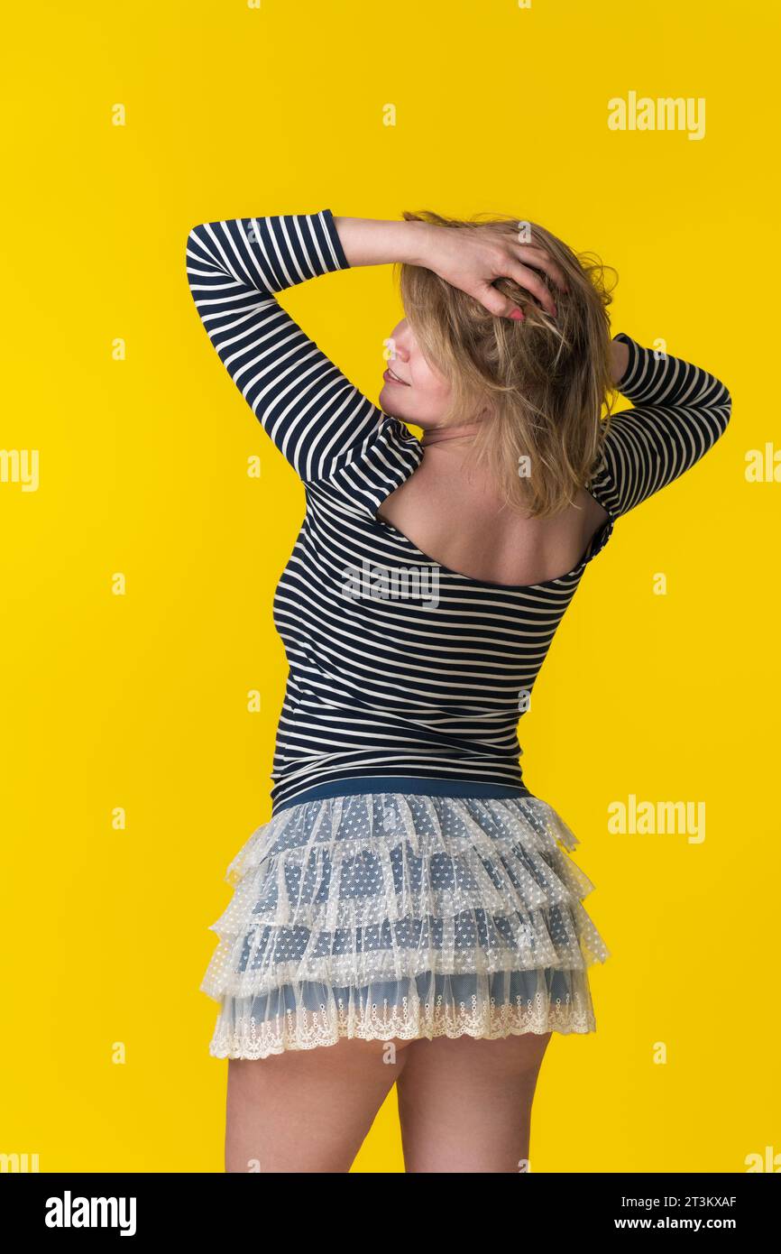 Mature blonde woman in striped long sleeve, miniskirt standing with hands behind head, obscured face Stock Photo