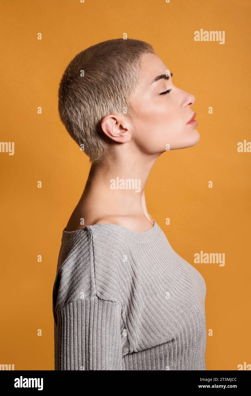 Profile of side view of thoughtful young female blonde with short hair and closed eyes standing in room with chin up against orange wall Stock Photo