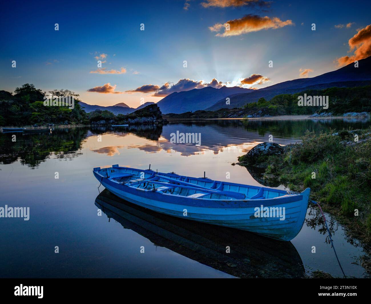 A blue fishing boat sits on the Upper Lake Killarney, with the sun setting over Macgillycuddy's Reeks, Killarney National Park, County Kerry, Ireland Stock Photo