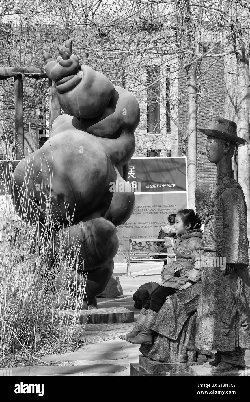 Black And White Photo Of A Young Chinese Girl Sitting By A Contemporary Sculpture In The 798 Art Zone, Dashanzi Art District, Beijing, China. Stock Photo