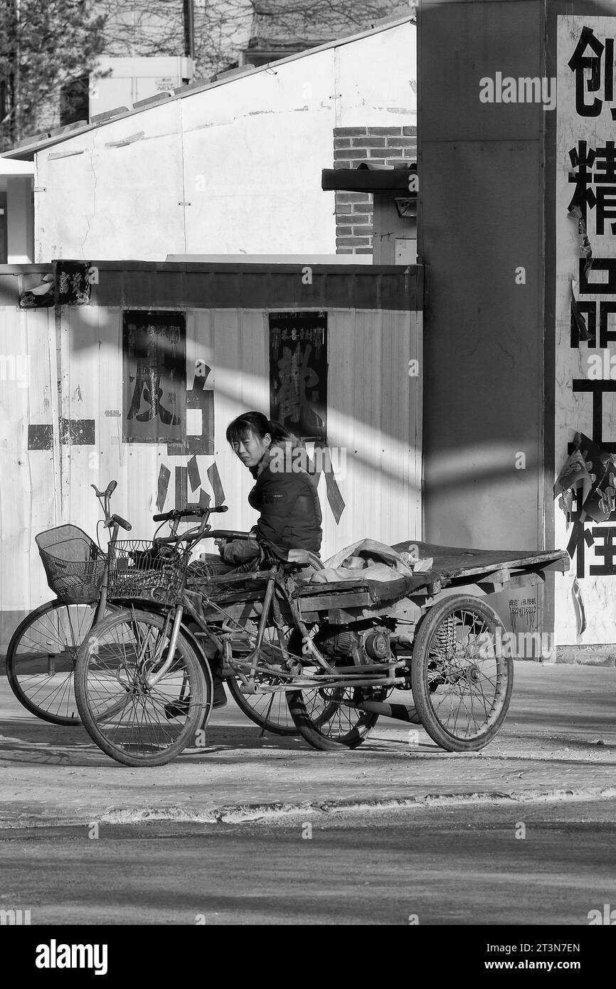 Black And White Photo Of A Young Chinese Woman Sitting On A Decrepit Auto Rickshaw In The 798 Art Zone (Dashanzi Art District), Beijing, China. Stock Photo