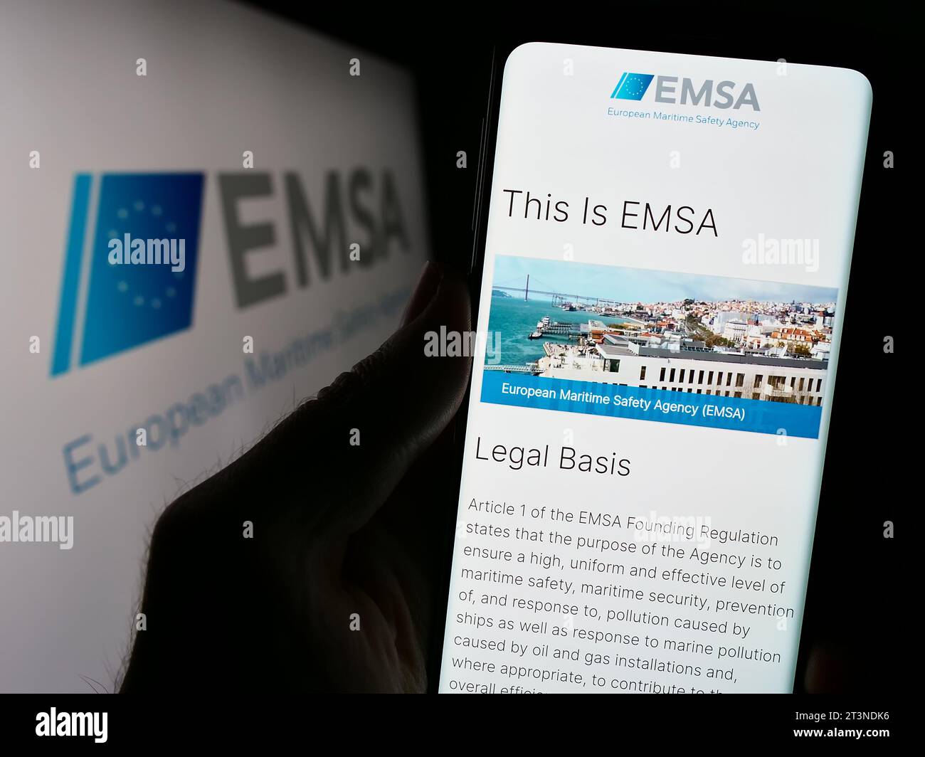 Person holding cellphone with webpage of EU institution European Maritime Safety Agency (EMSA) in front of logo. Focus on center of phone display. Stock Photo