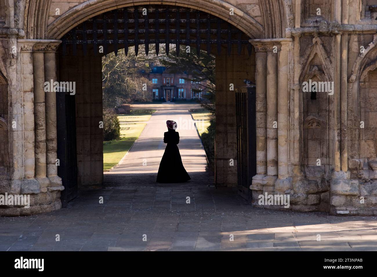 A woman wearing a hat and a full length victorian dress, standing at the entrance gate of a large English manor house Stock Photo