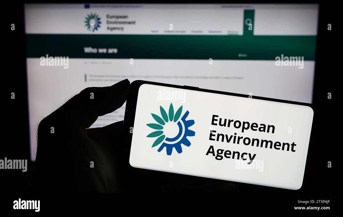 Person holding mobile phone with logo of EU institution European Environment Agency (EEA) in front of web page. Focus on phone display. Stock Photo