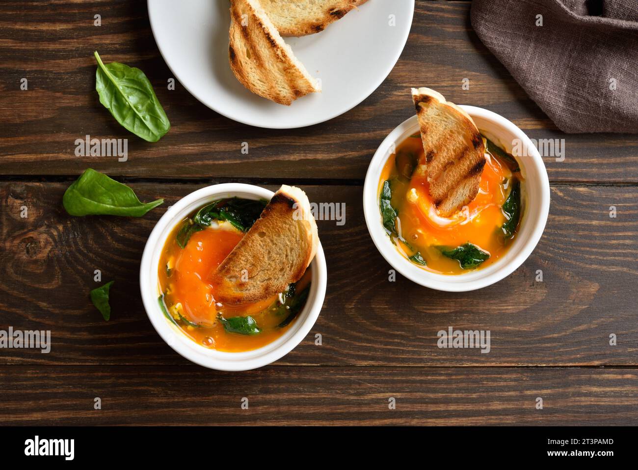 Baked eggs(Eggs en cocotte) in ramekins with cream, сheese and spinach on wooden background. Served with warm toast for dipping. Top view, flat lay Stock Photo