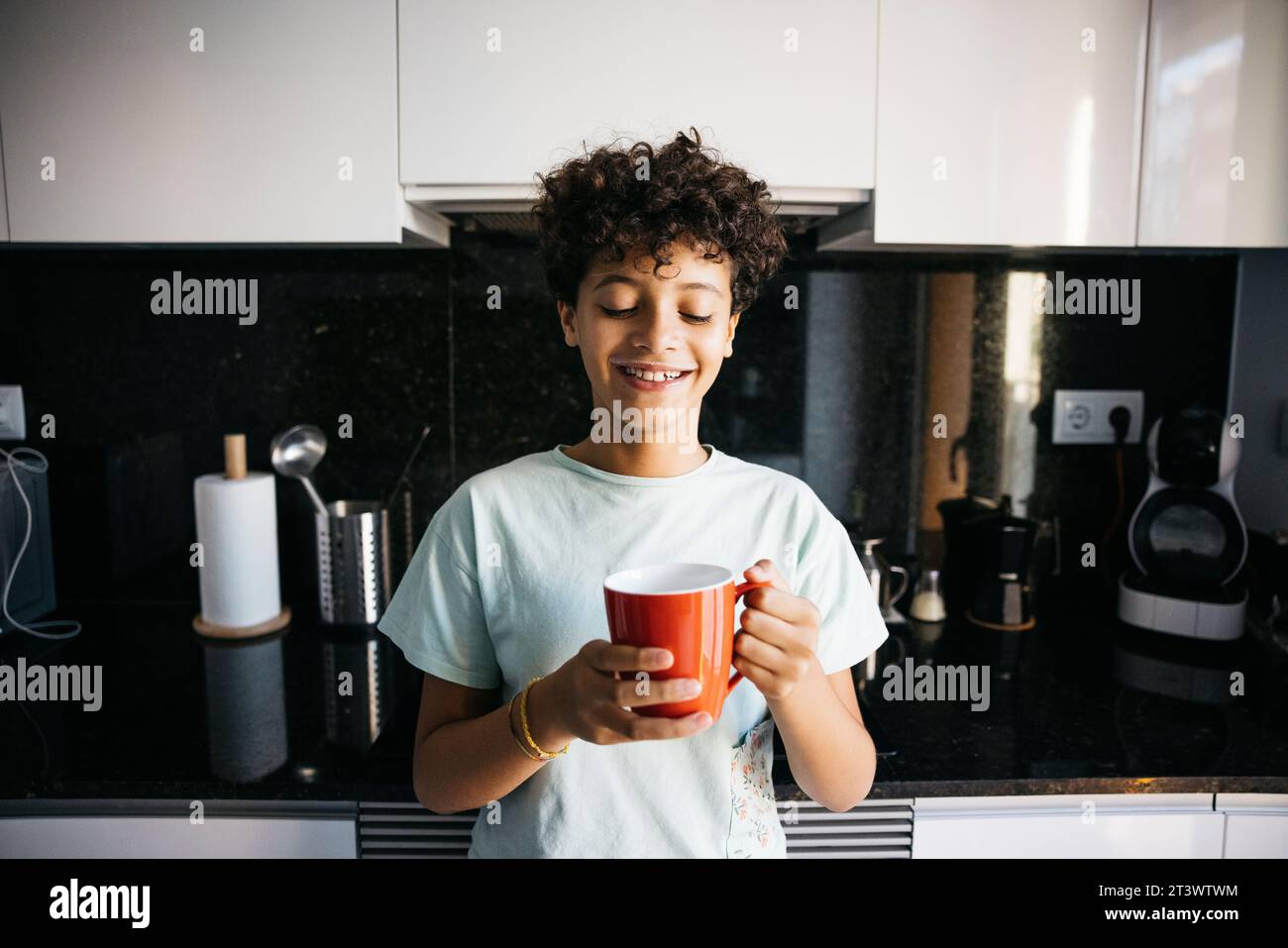 Smiling ten years old girl drinking milk from a red mug in the kitchen. Girl at home drinking a cup of milk. Stock Photo