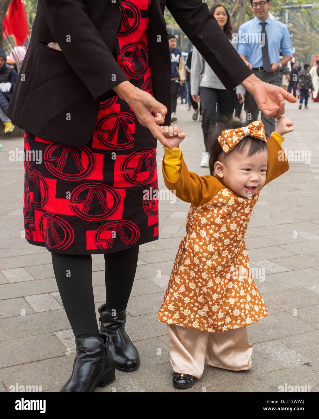 A Vietnamese girl toddler wearing traditional ao dai smiles as her mother holds her hands and she walks at Tet in Hanoi, Vietnam. Stock Photo
