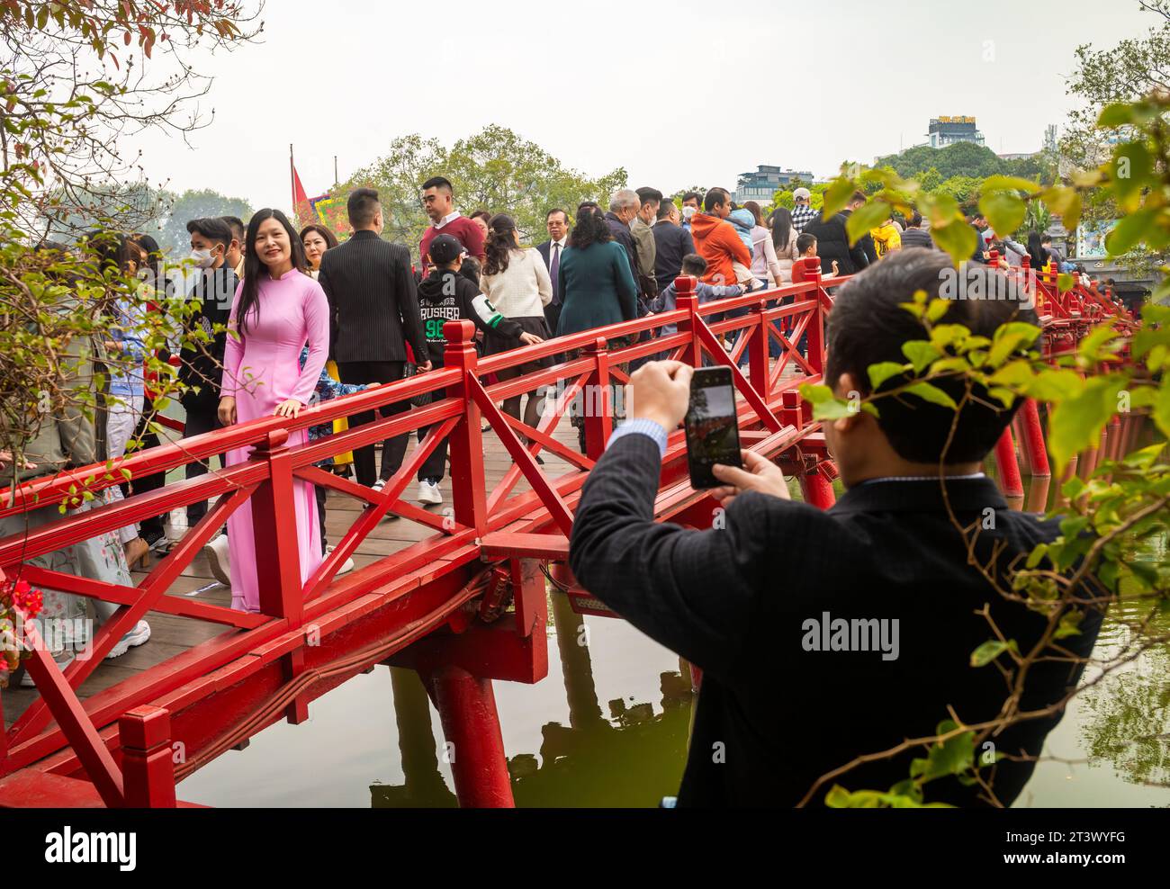 A middle-aged Vietnamese woman wearing a pink ao dai has her photo taken by her husband on The Huc bridge leading to Ngoc Son Temple on Hoan Kiem lake Stock Photo