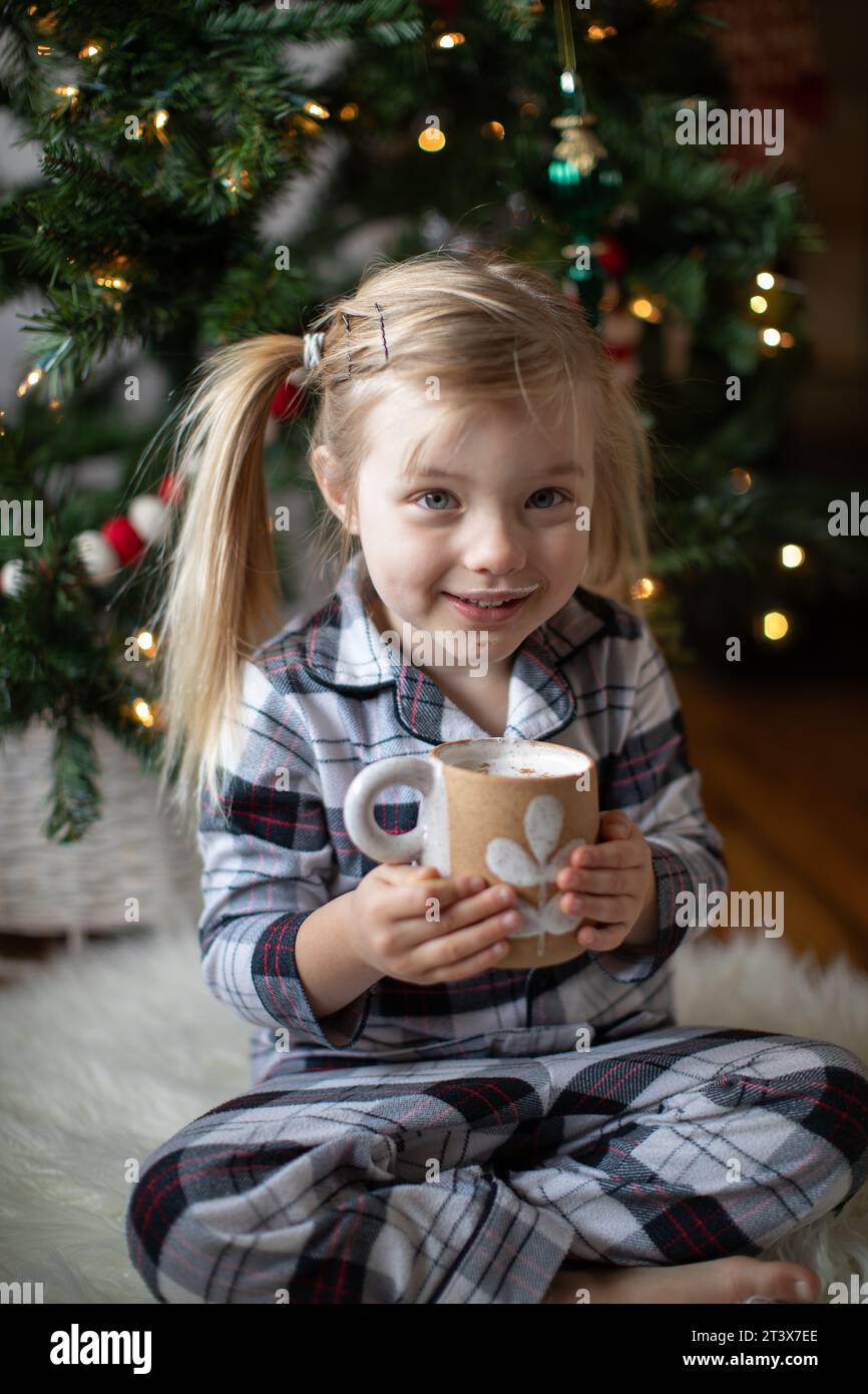 Happy toddler enjoys cocoa in her pjs, next to Christmas tree Stock Photo