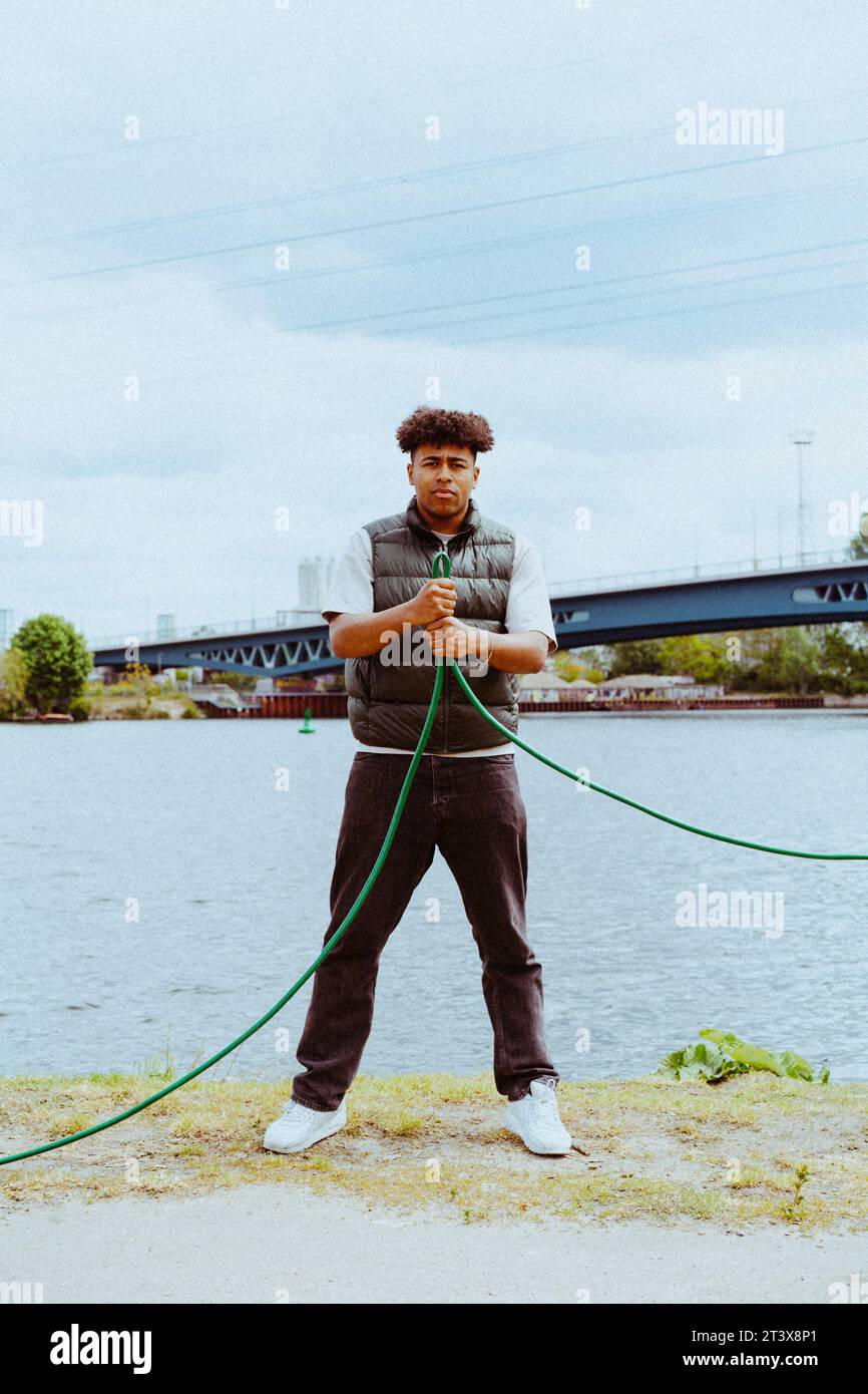 Full length portrait of confident young man holding garden hose near river Stock Photo