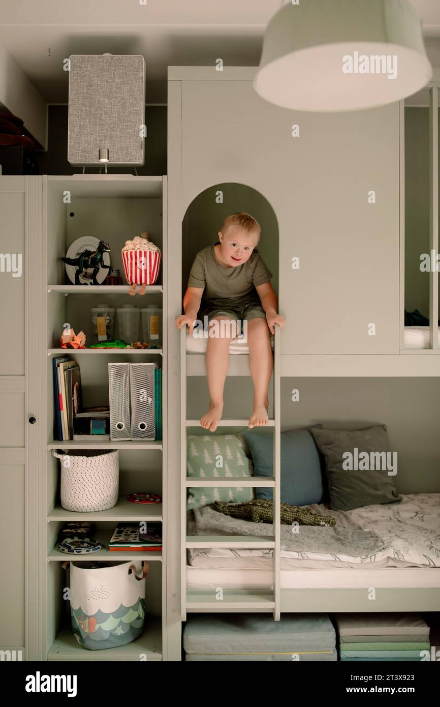 Full length portrait of boy with down syndrome sitting on bunkbed ladder at home Stock Photo