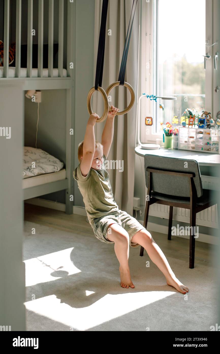 Full length of happy boy hanging from gymnastic rings in bedroom at home Stock Photo