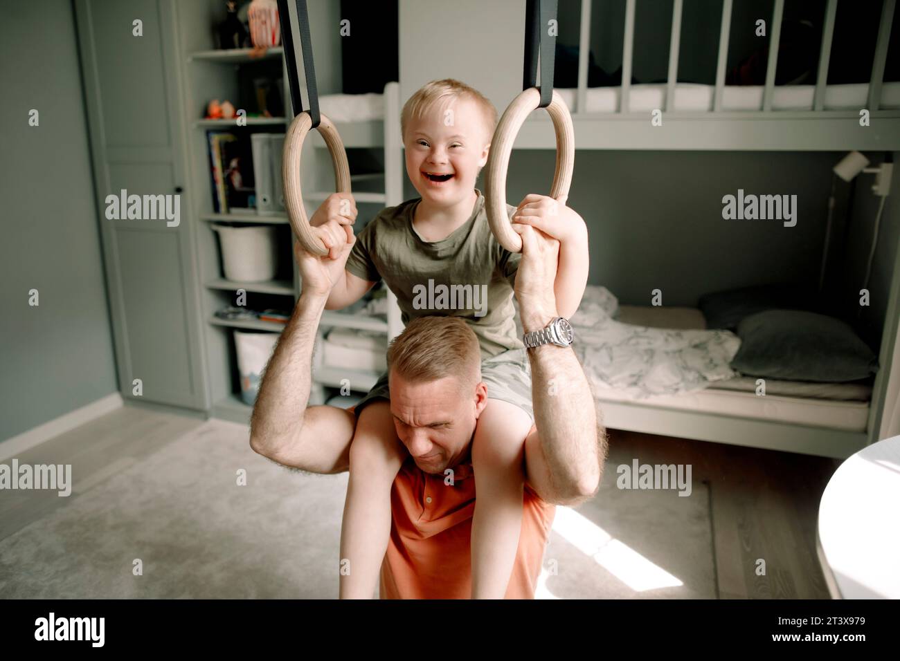 Father carrying son with down syndrome on shoulders while holding gymnastic rings in bedroom Stock Photo