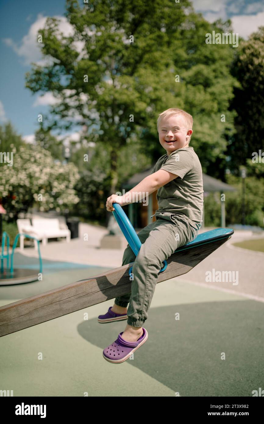 Full length side view portrait of happy boy with down syndrome sitting on seesaw at park Stock Photo