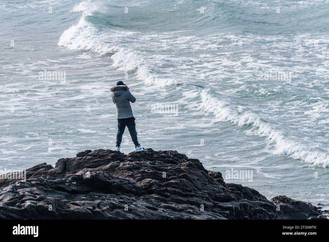 A holidaymaker standing on rocks watching incoming waves at Fistral in Newquay in Cornwall in the UK. Stock Photo