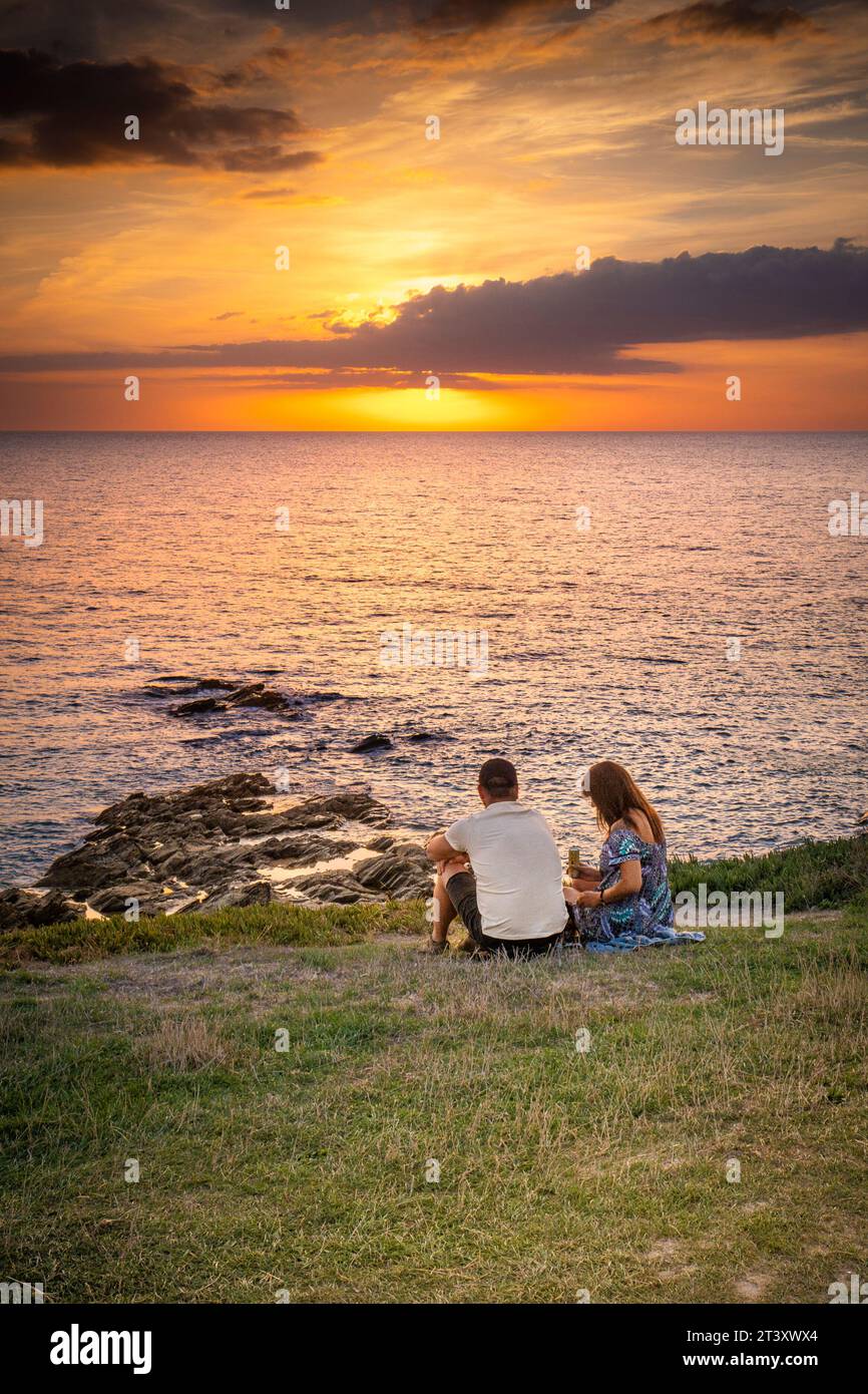 Holidaymakers sitting on the coast enjoying a spectacular sunset over Fistral Bay in Newquay in Cornwall in the UK in Europe. Stock Photo