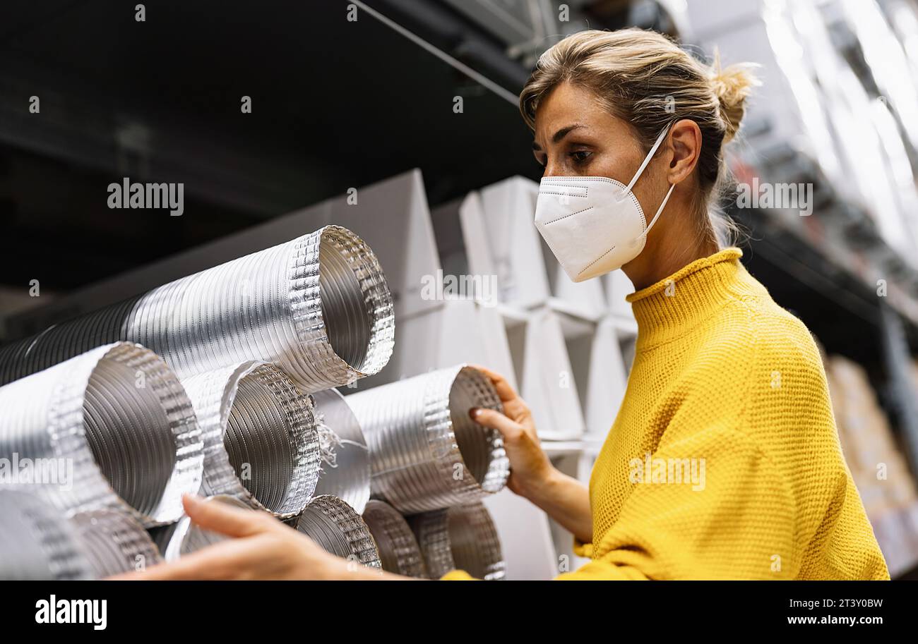 woman wearing a face mask and a yellow sweater examines silver ventilation ducts in a warehouse or store setting.  Corona safety Concept image Stock Photo