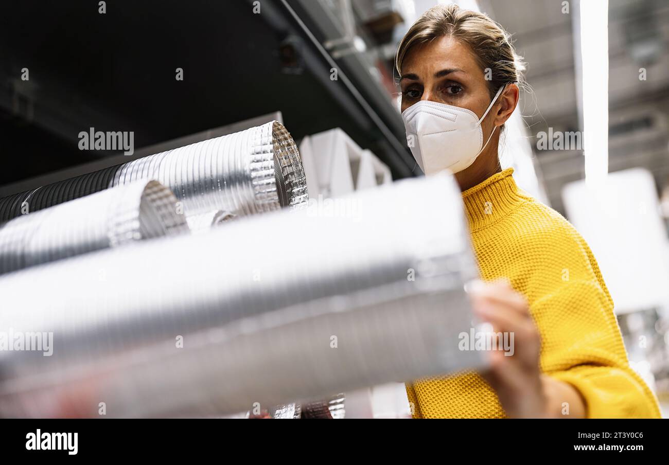 woman with face mask and a yellow sweater examines silver ventilation ducts in a warehouse or store setting. Corona safety Concept image Stock Photo