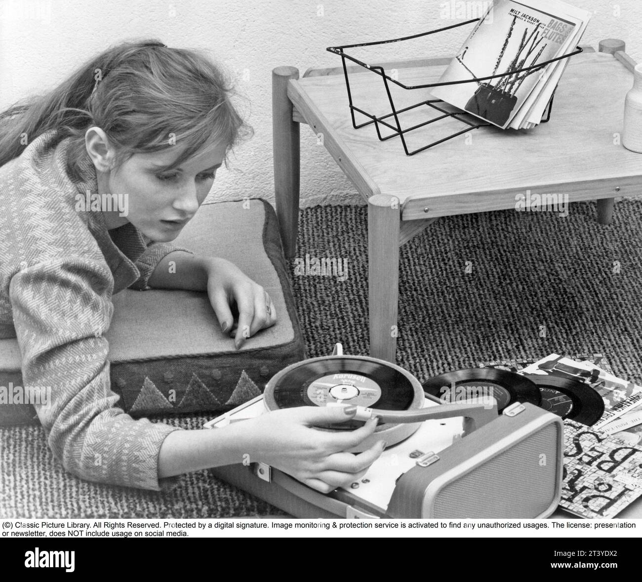 Woman at a gramophone in the 1950s. A typical 1950s teenage girl lying on the floor listening to music from a small gramophone. She is playing the type of records called singles. A record that typically contained a recording of fewer tracks than an LP record. This single record was played at the speed 45 rpm (revolutions per minute) and was 7 inches in diameter, 17,8 cm. The first single record was released 31 march 1949 by RCA Victor. Sweden 1959 Stock Photo